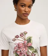 Remera blanca de algodón, corte slim fit, con cuello redondo y manga corta. Presenta un estampado bordado de flores rosadas y frutos con hojas verdes, acompañado de un pequeño texto en la parte inferior.