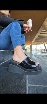 Zueco abierto de cuero charolado negro, con plataforma gruesa y taco ancho de 7.5 cm, con detalle texturizado y tachas en el borde de la suela.