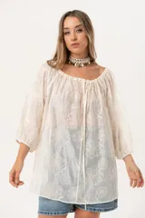Blusa oversize de color crudo con escote bote y lazo regulable. Confeccionada en tela liviana y traslúcida con bordado floral. Presenta mangas tres cuartos con puños elásticos.