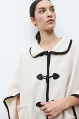 Capa color crudo con cuello camisero y ribetes negros. Cierre frontal con alamares negros y bolsillos laterales.