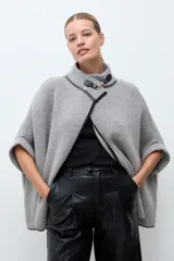 Capa de tejido de punto en color gris, con diseño abierto, cuello alto y cierre frontal mediante una hebilla metálica. Presenta bordes con ribete en color negro.