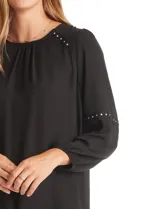 Blusa negra de gasa con cuello a la base fruncido, mangas largas amplias con puños elastizados y apliques de cinta con tachas en la parte delantera y antebrazos. Tiene una abertura central en la espalda con botón.