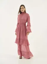 Vestido largo de gasa color rosa viejo, con cuello alto fruncido, cintura elástica tipo nido de abeja y mangas largas con volados. La falda presenta un diseño de capas con volados en el ruedo.