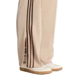 Pantalón deportivo Adidas de corte holgado, color beige con las tres tiras características en los laterales en tono marrón. Presenta diseño de pierna recta, bolsillos laterales y logo de la marca bordado.