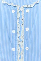 Chaqueta de popelín a rayas celestes y blancas, con cuello redondo y mangas largas. Presenta detalles de encaje a contraste en el cuello, los puños y la botonadura frontal, la cual cuenta con cierre de botones.