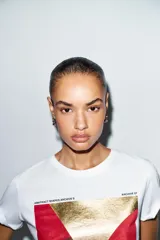 Camiseta de algodón color marrón oscuro, de corte regular, cuello redondo y manga corta. Presenta una estampación metalizada dorada en el parte frontal con texto "REYKJAVIK ART GALLERY" y "ABSTRACT MOVEMENTS Reykjavik, Iceland, 1975".