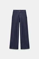 Jeans de tiro alto color azul oscuro, con cintura elástica en la espalda, bolsillos delanteros y bolsillos de plastrón en la espalda. Presenta pernera ancha y cierre frontal con cremallera y botón a presión.