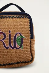 Mochila mini de mimbre sintético color tostado, con ribetes y asa superior de tela azul marino. Cuenta con bordado frontal con la palabra "Rio", un detalle de palmera y un tucán.