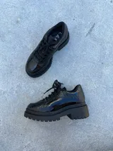 Zapato mocasín negro de cuero charol con diseño tipo reptil, cordones y suela track.