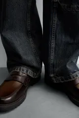 Mocasín de cuero marrón oscuro con antifaz en tono camel. Presenta horma redonda y suela track gruesa de goma negra.