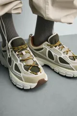 Zapatilla deportiva tipo running con diseño de suela voluminosa e irregular. Presenta una combinación de piezas en tonos beige y crema, con paneles de malla y cordones jaspeados.