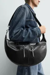 Bolso de hombro de diseño curvo en color negro, con detalle de pespuntes centrales. Cuenta con un compartimento principal con cierre de cremallera y dos bolsillos frontales con cierre. Incluye asa de hombro y bolsillo interior con cierre.