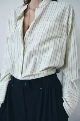 Camisa de corte oversize confeccionada en tejido con viscosa, con estampado de rayas verticales. Presenta cuello mao, cierre frontal con botones ocultos bajo solapa, mangas largas con puños anchos y bolsillos de plastrón en el delantero. El bajo es asimétrico y redondeado.