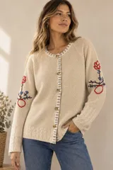 Saco de punto color beige, confeccionado en algodón, con cuello redondo, mangas largas y cierre frontal con botones. Presenta detalles bordados con diseño floral en las mangas y ribetes con rayas azules y blancas en la botonera y el cuello.