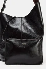 Bolso de hombro tipo saca asimétrica, color negro, con acabado de piel brillante. Posee un asa de hombro ajustable y cierre magnético. Incluye una cartera interior extraíble.