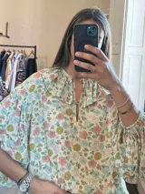 Camisa de manga larga con estampado floral multicolor sobre fondo beige, cuello redondo con volados y abertura frontal con tiras para anudar.