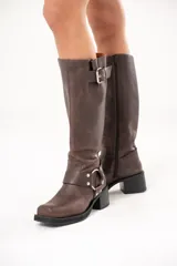 Bota biker de caña alta color marrón, confeccionada en cuero. Presenta hebilla en la parte superior, detalle de aro en la parte inferior y cierre lateral.