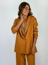 Blazer de mujer color caramelo con acabado metalizado y textura brillante. Diseño de solapas amplias, manga larga y un solo botón frontal. Se muestra como parte de un conjunto con pantalón a juego.