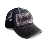 Gorra tipo trucker de color negro con panel frontal de tela desgastada y parte trasera de malla. Presenta un parche rectangular cosido con hilo blanco que incluye un estampado de estilo camuflaje y tipografía gótica. El diseño cuenta con detalles de roturas y efecto envejecido.