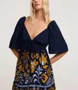 Vestido corto de viscosa con estampado floral y de follaje en tonos mostaza, blanco y azul marino. Presenta escote pronunciado en V con lazo y fruncido en el busto, mangas cortas abullonadas y elástico (lastex) en la espalda. La falda es evasé con un ruedo con volado.