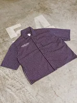 Camisa de manga corta con corte híbrido boxy fit/oversize, confeccionada en algodón. Disponible en tres diseños: a cuadros marrones, a rayas blancas y negras, y a rayas burdeos y blancas. Presenta bordados de diseño propio de la marca.