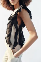 Blusa de cuello redondo y manga sisa, de color beige con rayas verticales. Presenta volantes negros en las sisas y una tira de encaje central con jaretas. Cuenta con cierre en la espalda con abertura y botón.