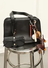 Estuche para lentes de cuero sintético negro con textura granulada y forma irregular. Tiene un cierre con broches dorados y una correa delgada con gancho para colgar de la cartera.