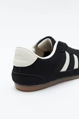 Zapatilla urbana de perfil bajo con diseño retro, confeccionada en poliuretano con detalles en textura tipo gamuza. Presenta puntera redondeada, cordones clásicos y suela de goma color caramelo.