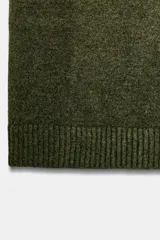 Chaleco de punto en color verde oliva, con escote en pico y sisas amplias. Presenta terminaciones acanaladas en el cuello, las sisas y el ruedo.