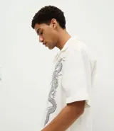 Camisa de manga corta con cuello cubano, confeccionada en sarga de algodón. Presenta un corte boxy holgado y un diseño estampado con dragones en ambos lados del frente.