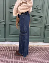 Pantalón tipo jean cargo de corte ancho y tiro alto, confeccionado en denim azul oscuro. Presenta bolsillos laterales tipo cargo con solapa y costuras reforzadas a lo largo de las piernas.