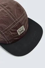 Gorra de visera plana, color marrón oscuro, confeccionada en tejido técnico con costuras contrastantes en blanco. Presenta un parche frontal con un diseño a cuadros y texto.