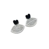 Par de aros colgantes con forma de labios delineados en blanco y rellenos de glitter plateado, con un corazón negro en la parte superior.