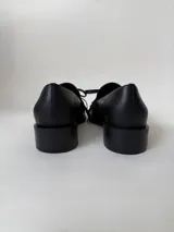 Mocasín de cuero marrón con textura granulada, diseño de horma cuadrada, detalle de flecos y lazo decorativo en el empeine, montado sobre una suela y taco bajo de color negro.