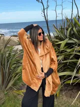Campera oversize de abrigo color camel en tejido suave con efecto gamuzado, con gran capucha envolvente que puede usarse como bufanda, bolsillos laterales y puños elastizados negros.