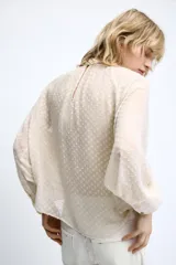 Camisa cruda de manga larga con textura plumeti, cuello redondo y lazada en el mismo tejido. Cuenta con mangas abullonadas terminadas en puños anchos y cierre en la espalda con abertura y botones.
