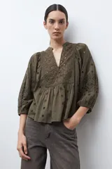 Blusa color verde oliva de manga 3/4 con volumen, cuello mao con abertura en V y botones. Presenta bordados y detalles de broderie calado en el canesú frontal y en las mangas. El cuerpo tiene un corte peplum con frunces.