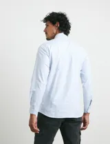 Camisa de manga larga con estampado de rayas verticales finas en color azul y blanco. Presenta cuello button-down, cierre frontal con botones nacarados y un bolsillo en el pecho.