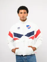 Buzo deportivo de nylon con medio cierre frontal y cuello alto. Presenta un diseño de bloques de color en azul, blanco y bordó, con el escudo del Club Nacional de Football y el logo de Umbro en el pecho.