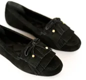 Mocasín de gamuza color negro con detalle de flecos y lazo frontal con herrajes metálicos dorados. Cuenta con interior forrado en cuero y suela de goma flexible.