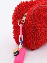 Necessaire de textura tipo felpa o chenille en color celeste, con cierre perimetral y etiqueta frontal de la marca Common Wear. Incluye un detalle de cadena plástica multicolor con una placa rosa colgante en el lateral.