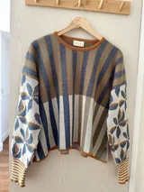Sweater de tejido de punto con silueta oversize, cuello redondo y mangas largas abullonadas. Presenta un diseño patchwork con rayas verticales en tonos rojo, rosa y gris verdoso en el cuerpo, y un estampado floral en tonos rosa y rojo con detalles dorados en las mangas. Los puños y el cuello tienen un ribete a rayas horizontales.