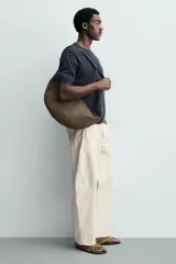 Bolso de hombro con diseño de media luna, confeccionado en gamuza color marrón. Cuenta con un compartimento principal con cierre de cremallera y un bolsillo interior con cierre. Posee un asa de hombro integrada.