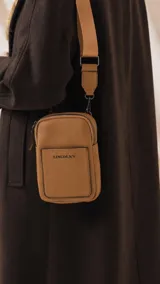 Bandolera pequeña de diseño compacto en color mostaza, confeccionada en material sintético con textura granulada. Presenta un bolsillo frontal con el logo de la marca impreso, cierre superior y correa ancha ajustable y desmontable.