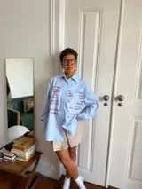 Camisa oversize de corte recto, confeccionada en tela de algodón a rayas verticales celestes y blancas. Presenta cuello clásico, cierre frontal con botones y mangas largas. El diseño destaca por una estampa frontal con palabras relacionadas al amor en diversos idiomas en color bordó.