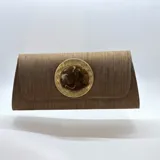 Bolso rígido alargado de tela marrón con solapa y aplique central de piedra de resina sobre una base metálica dorada circular. Incluye asa de cadena desmontable.