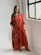 Vestido largo color terracota con diseño asimétrico que incluye un lateral con estampado floral grande. Presenta mangas tres cuartos, escote redondo y un bolsillo aplicado en el frente.