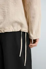 Camisa de manga larga color beige claro con textura jacquard arrugada, cuello solapa con abertura en V y bolsillo canguro frontal. Posee cordones ajustables en el bajo lateral.