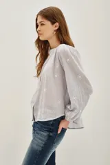 Camisa de algodón color celeste claro, con textura sutil y pequeños bordados blancos en forma de rombos. Presenta cuello mao, botones frontales y mangas largas abullonadas con puños ajustados.