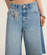 Bermuda tipo jorts de tiro medio, confeccionada en denim de algodón color azul claro. Presenta un corte holgado que llega por debajo de la rodilla, con trabillas en la cintura, bolsillos laterales y traseros, y un detalle de cordón decorativo en la parte frontal.